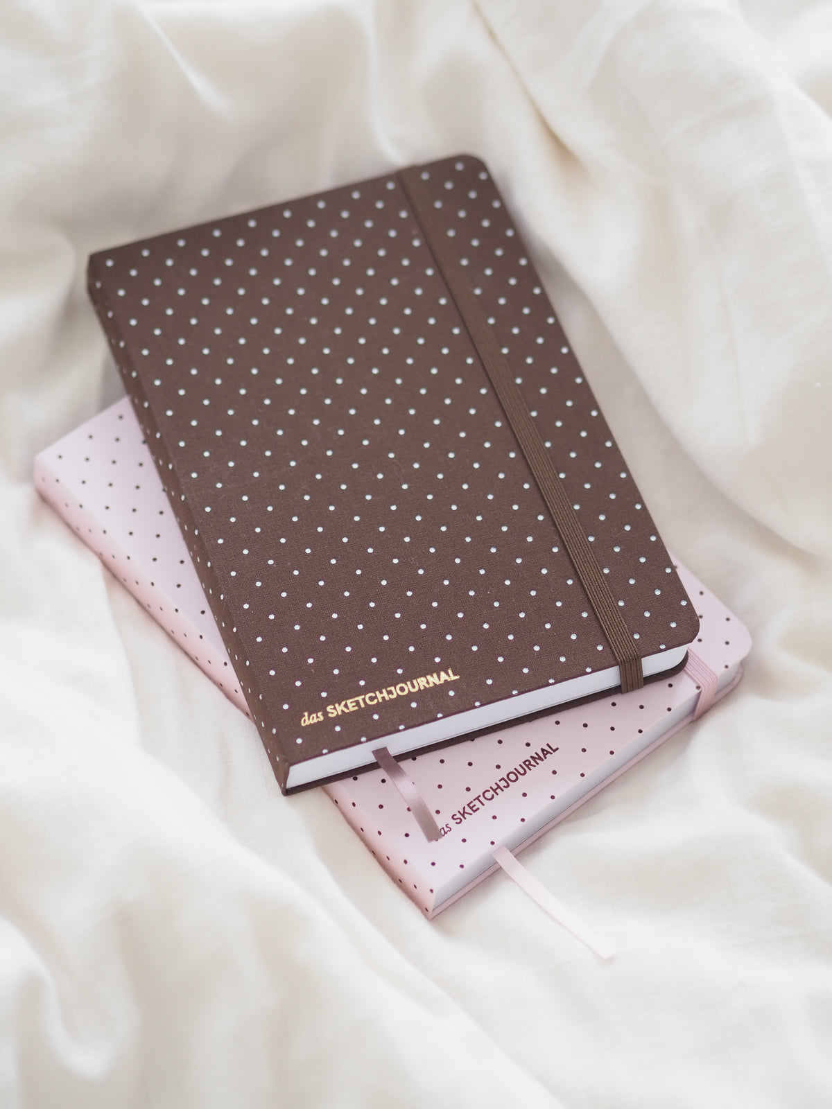 Braunes und rosa Sketchjournal mit Polka-Dots und Lesebändchen auf weißer Bettdecke
